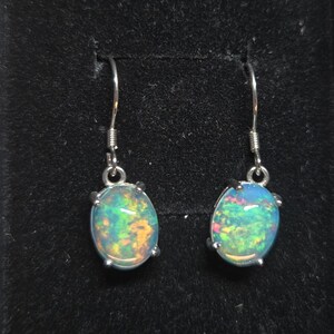 Pu&ograve; includere: Un paio di orecchini pendenti con opali di forma ovale incastonati in griffe di metallo color argento. Ogni opale mostra un vivace gioco di colori con riflessi verdi, arancioni e blu. Gli orecchini sono fissati a ganci in metallo color argento.