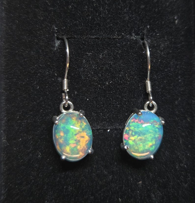 Pu&ograve; includere: Un paio di orecchini pendenti con opali di forma ovale incastonati in griffe di metallo color argento. Ogni opale mostra un vivace gioco di colori con riflessi verdi, arancioni e blu. Gli orecchini sono fissati a ganci in metallo color argento.