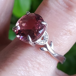 May include: A silver ring with a large, round, pink gemstone set in a floral design. The ring has a delicate, leafy band.