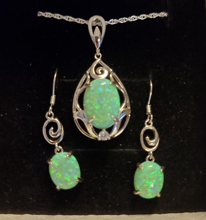 May include: A silver necklace and earrings set with a teardrop-shaped pendant and matching earrings. Each piece features a green opal stone surrounded by a delicate silver design.