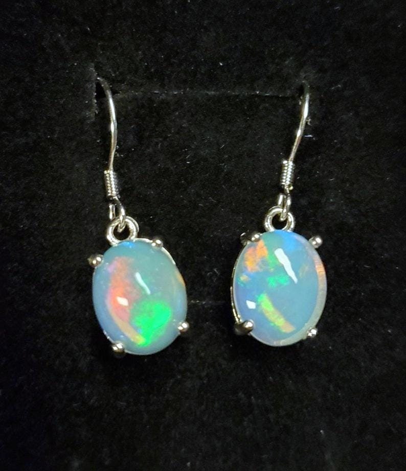 May include: Pair of elegant sterling silver opal earrings. Each earring features a light blue oval-shaped opal gemstone with vibrant internal flashes of orange, green, and blue. The opals are securely set in silver claw settings, adding to their refined look. These dangle earrings are perfect for adding a touch of sophistication to any outfit.