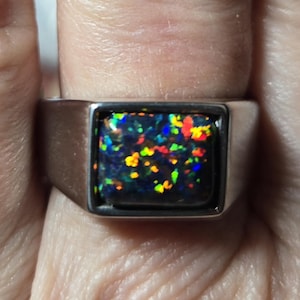 May include: A silver-toned ring featuring a rectangular setting with a dark, iridescent stone. The stone displays a vibrant play of color with flecks of red, green, yellow, and blue against a deep black background, set within a smooth metal band.