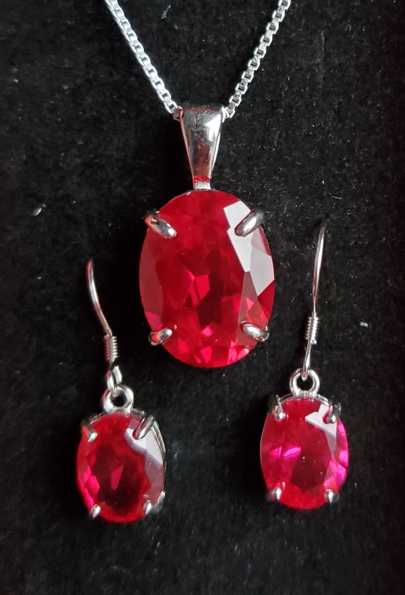 Puede incluir: Un conjunto de collar y pendientes de plata con piedras preciosas ovaladas rojas. El collar tiene una cadena delicada y un colgante con una piedra roja grande. Los pendientes son simples tachuelas con piedras rojas m&aacute;s peque&ntilde;as.