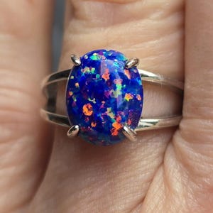 May include: A silver ring featuring a large, oval-shaped opal. The opal is a deep blue with vibrant flashes of orange, yellow, and green. The ring has a split shank design, with the opal held in place by four prongs.