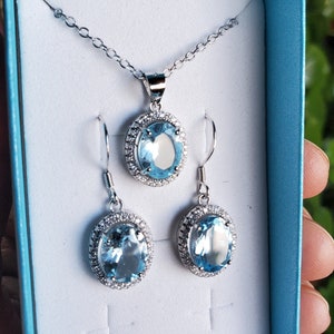 May include: A silver-toned jewelry set featuring a pendant necklace and matching earrings. Each piece showcases a large, oval, light blue gemstone surrounded by a halo of smaller, clear stones. The set is presented in a light blue box.