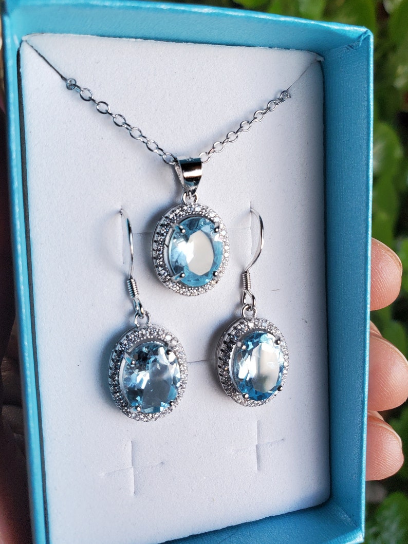May include: A silver-toned jewelry set featuring a pendant necklace and matching earrings. Each piece showcases a large, oval, light blue gemstone surrounded by a halo of smaller, clear stones. The set is presented in a light blue box.