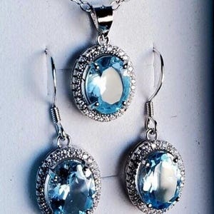 May include: A silver necklace with a matching pendant and a pair of earrings. Each piece features a large, oval, light blue gemstone surrounded by a halo of smaller, clear stones. The jewelry set is displayed against a white background.