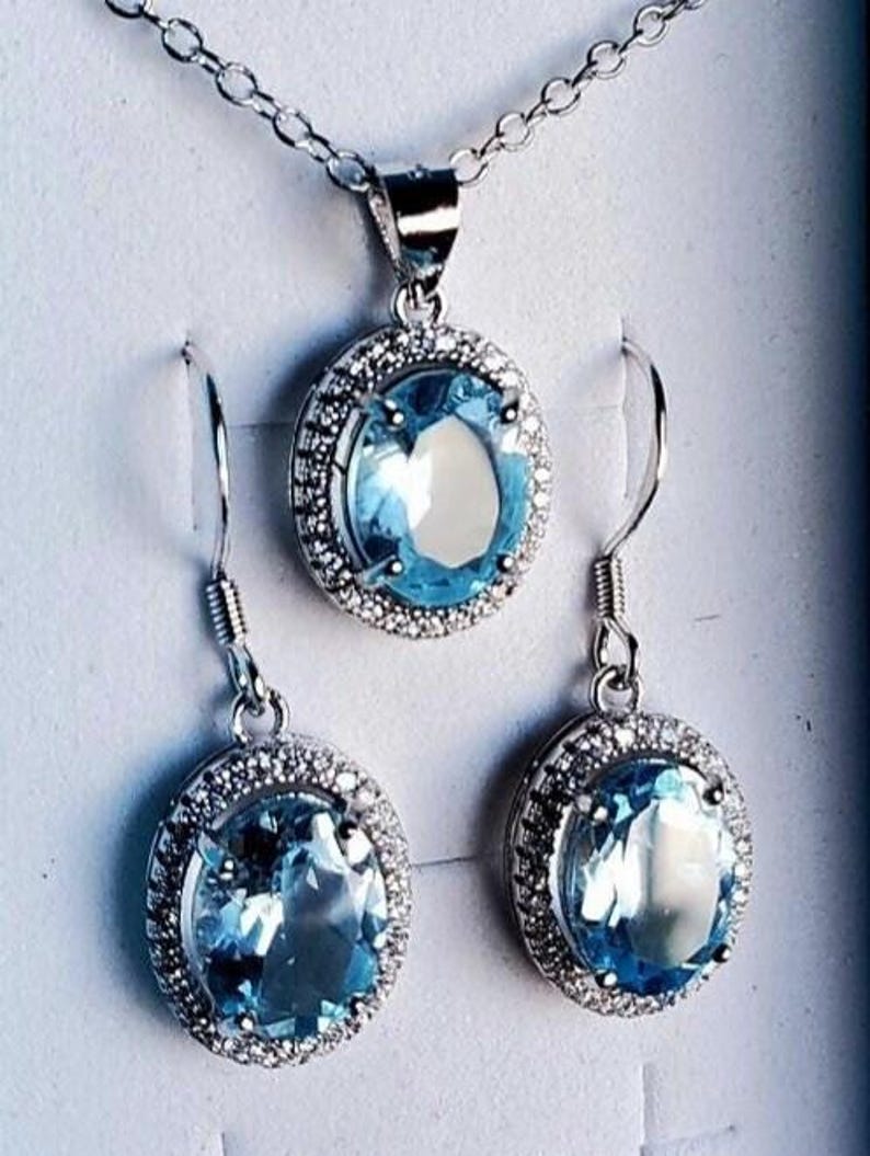 May include: A silver necklace with a matching pendant and a pair of earrings. Each piece features a large, oval, light blue gemstone surrounded by a halo of smaller, clear stones. The jewelry set is displayed against a white background.