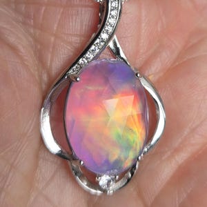 May include: A silver pendant necklace featuring an oval, faceted opal gemstone. The opal displays a vibrant play of color, with hues of pink, orange, yellow, and green. The pendant is accented with small, clear gemstones.