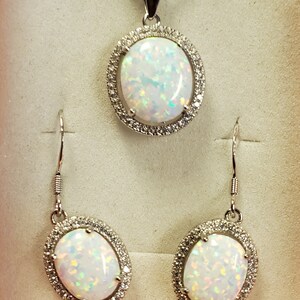 May include: A silver necklace and earrings set with oval-shaped white opal stones surrounded by a halo of smaller clear stones.