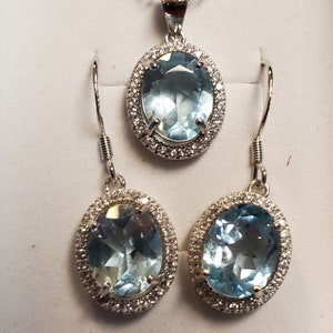May include: A jewelry set featuring a pendant necklace and matching earrings. The set includes an oval-shaped, light blue gemstone pendant and earrings, each surrounded by a halo of small, clear stones. The jewelry is set in a silver-colored metal.