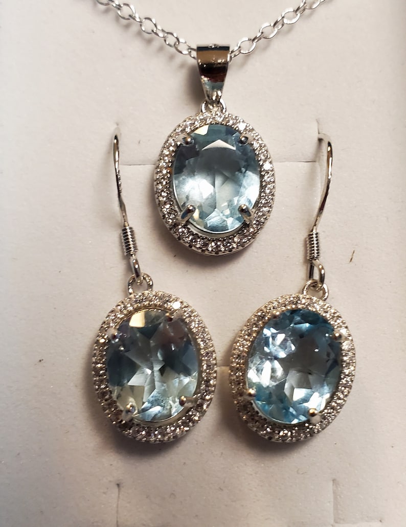 May include: A jewelry set featuring a pendant necklace and matching earrings. The set includes an oval-shaped, light blue gemstone pendant and earrings, each surrounded by a halo of small, clear stones. The jewelry is set in a silver-colored metal.