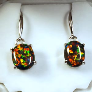 May include: A pair of silver drop earrings featuring oval-shaped black opals with vibrant flashes of color. The opals display a mix of orange, yellow, green, and blue hues. The earrings have a simple, elegant design, perfect for everyday wear.