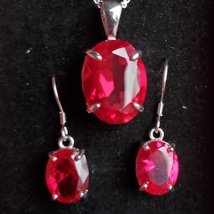 Puede incluir: Un collar de plata con un colgante ovalado rojo y pendientes a juego. El colgante y los pendientes presentan una gran piedra roja facetada.
