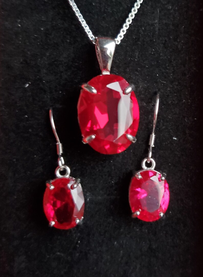 Puede incluir: Un collar de plata con un colgante ovalado rojo y pendientes a juego. El colgante y los pendientes presentan una gran piedra roja facetada.