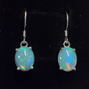 Pu&ograve; includere: Un paio di orecchini pendenti con pietre cabochon ovali e traslucide, caratterizzate da riflessi iridescenti blu, verdi e arancioni. Le pietre sono montate su griffe in metallo argentato e fissate a semplici ganci per orecchie in metallo.