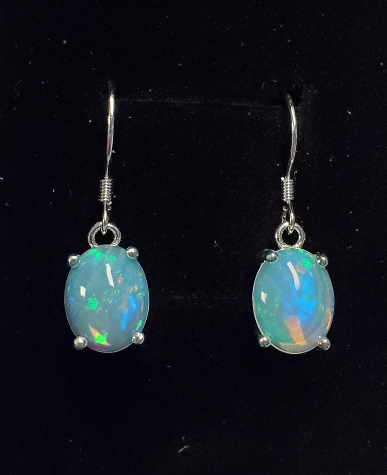Pu&ograve; includere: Un paio di orecchini pendenti con pietre cabochon ovali e traslucide, caratterizzate da riflessi iridescenti blu, verdi e arancioni. Le pietre sono montate su griffe in metallo argentato e fissate a semplici ganci per orecchie in metallo.