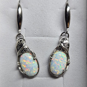 May include: A pair of silver-toned drop earrings. Each earring features an oval, iridescent opal stone set in a decorative silver frame. The earrings have a hook closure and are displayed against a gray background.