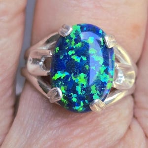 May include: Oval-shaped ring featuring a vibrant blue opal with green flecks, set in a silver-toned band. The opal's surface reflects light, creating a shimmering effect. The ring's design is simple yet elegant, suitable for everyday wear.