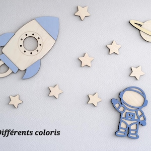 Peut inclure: Décorations murales en bois sur le thème de l'espace, comprenant une fusée, un astronaute, une planète avec des anneaux et des étoiles. La fusée et l'astronaute sont soulignés en bleu. Le texte "Différents coloris" est en bas.