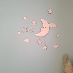 May include: Wooden wall decor featuring a crescent moon with closed eyes, stars, clouds, and the name "Eliott" in cursive. The light-colored pieces are mounted on a light blue wall, creating a whimsical nursery theme.