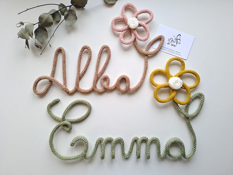 May include: Two names, Alba and Emma, crafted from knitted yarn in light pink and sage green. The name Alba is attached to a pink flower with a white center, and the name Emma is attached to a mustard yellow flower with a white center. A small white card with the text L'&egrave;re de rien is included.