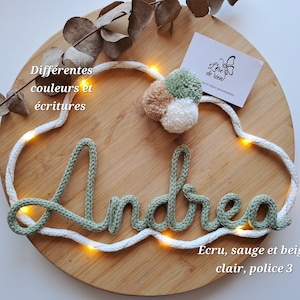 May include: A decorative wall hanging featuring the name Andrea in sage green knitted yarn, shaped into a cloud outline with integrated warm white LED fairy lights. The piece includes a cluster of three small pom-poms in sage, beige, and white.