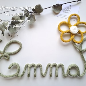 May include: A sage green knitted cord shaped into the name Emma, featuring a mustard yellow knitted flower with a white center attached to the end. The text Diff&eacute;rents mod&egrave;les et coloris, Sauge, moutarde et &eacute;cru, police 1 is visible.