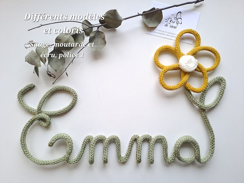 May include: A sage green knitted cord shaped into the name Emma, featuring a mustard yellow knitted flower with a white center attached to the end. The text Diff&eacute;rents mod&egrave;les et coloris, Sauge, moutarde et &eacute;cru, police 1 is visible.