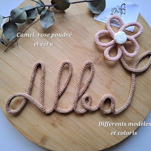 May include: A camel-colored knitted cord shaped into the name Alba in cursive script, accented with a light pink six-petaled flower featuring a white center. The text "Camel, rose poudre et ecru" and "Differents modeles et coloris" is visible.