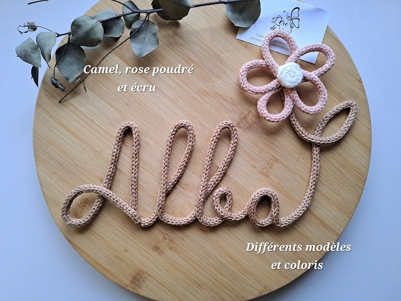 May include: A camel-colored knitted cord shaped into the name Alba in cursive script, accented with a light pink six-petaled flower featuring a white center. The text "Camel, rose poudre et ecru" and "Differents modeles et coloris" is visible.