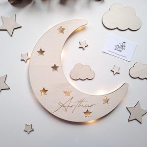 Può includere: Luce notturna a forma di luna in legno con ritagli a forma di stella e il nome "Arthur" inciso. Include decorazioni in legno a forma di stella e nuvola. La luna ha un bagliore caldo e soffice. È presente anche una piccola carta con un logo.
