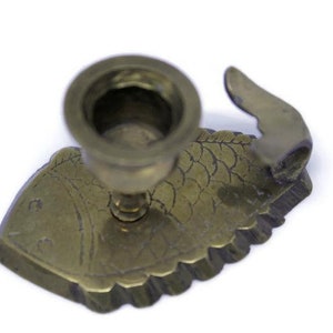 May include: A brass candle holder in the shape of a fish. The candle holder has a round base and a fish-shaped body with scale detailing. The fish has a fin on its back and is set against a white background.