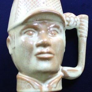 May include: A ceramic mug shaped like a person's head wearing a hat. The mug is light green and brown with a textured surface. The person has a serious expression and is wearing a hat with a grid pattern.