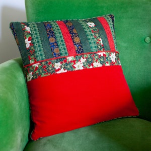 Peut inclure: Un coussin carré au design patchwork. La partie supérieure présente des bandes verticales de tissu vert, rouge et bleu avec des motifs floraux et géométriques. Une bande horizontale à motif de poinsettias sépare le haut d'une base rouge unie.
