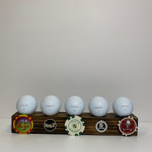 May include: Six white golf balls with black lettering on a wooden display stand. The golf balls are labeled "Titleist" and have numbers 1 and 4 on them. The stand has five circular plaques with different designs and text. The plaques include "1 Putt", "Palm Valley Golf Club", "Country Club of the South", "East Lake 1904", and "Gambling Oaks Glutton".