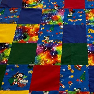 Könnte beinhalten: Ein Patchwork-Quilt mit einem Gittermuster aus farbigen Quadraten. Einfarbige Quadrate in Rot, Gelb, Grün und Blau werden mit blauen Quadraten kombiniert, die mit Mickey Mouse und einem Galaxiemuster bedruckt sind. Der Quilt liegt auf einer weißen Tagesdecke.
