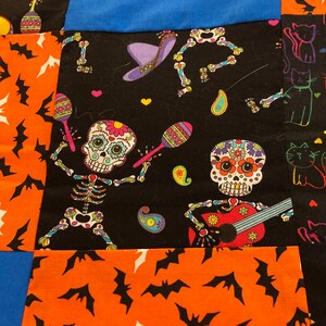 Peut inclure: Un textile patchwork avec des motifs sur le thème du Jour des Morts et d'Halloween. Le tissu comprend des panneaux orange avec des chauves-souris noires, des panneaux noirs avec des squelettes de crânes de sucre jouant des instruments et des panneaux bleus.