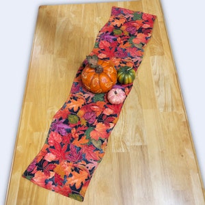 May include: A fall-themed table runner with a dark background and colorful autumn leaves. The runner is decorated with three small pumpkins.