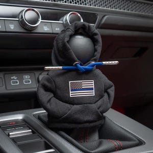 Può includere: Una felpa con cappuccio nera con un design di bandiera americana a linea blu sottile, attaccata a un pomello del cambio in un'auto.