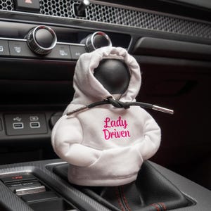 Könnte beinhalten: Ein weißer Hoodie-Schalthebel mit dem Text "Lady Driven" in rosa Schrift.
