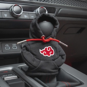 May include: A black fabric gear shifter cover designed as a miniature hoodie. The hoodie features a red and white cloud graphic on the front chest and includes bright red drawstrings with silver metal aglets at the ends of the strings.