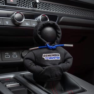 May include: A black hooded figure with a blue drawstring and a white sticker that says "Remember the Buster" attached to the chest. The figure is sitting in a car's gear shift.