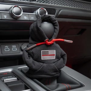 May include: Black hooded gear shifter with a red drawstring and a thin red line American flag patch. The gear shifter is in a car.