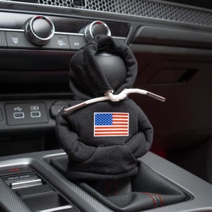 May include: A black hoodie gear shifter with a white drawstring and an American flag patch. The gear shifter is in a car's gear shift console.