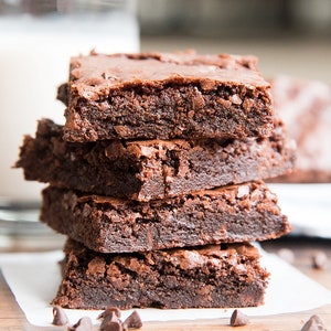 May include: A stack of four fudgy chocolate brownies, with a few chocolate chips scattered on a white surface. The brownies are dark brown and have a slightly cracked texture.