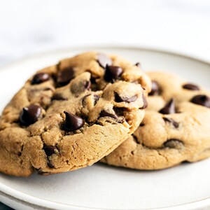 May include: Two chocolate chip cookies on a white plate. The cookies are golden brown and have a soft, chewy texture. The chocolate chips are melted and gooey.