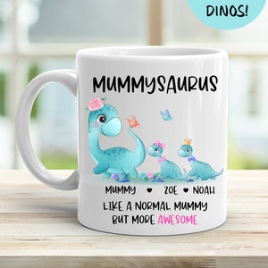 May include: White ceramic mug featuring a blue dinosaur illustration with the text "Mummyosaurus" and "Like a normal mummy but more awesome". The dinosaur has a pink flower crown and is holding a butterfly. Two smaller dinosaurs are behind the larger dinosaur. The text "Mummy", "Zoe", and "Noah" are below the dinosaurs.