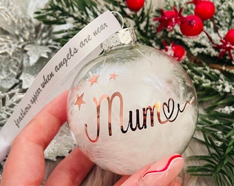 May include: Clear glass Christmas ornament filled with white feathers and the word "Mum" in rose gold script, accented with stars. A white ribbon with the text "feathers appear when angels are near" hangs from the ornament.