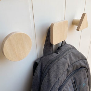 Puede incluir: Tres ganchos de pared de madera en forma de círculo, cuadrado y triángulo. Los ganchos están montados en una pared blanca. Una mochila gris está colgada del gancho cuadrado.